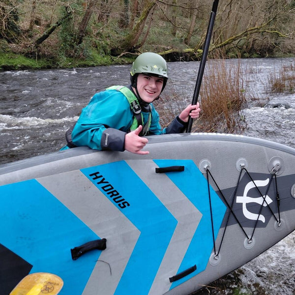 A young paddler in a drysuit and helmet smiling with a thumbs up on a river with a Hydrus board