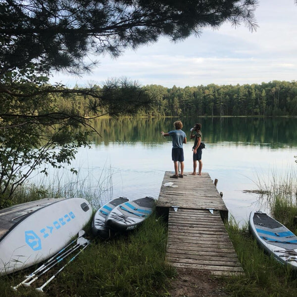 Two paddlers on a wooden dock at a forest lake with Hydrus boards on the shore