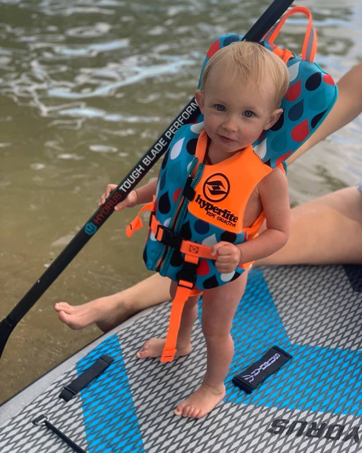 Paddler on an inflatable paddleboard wearing a snug-fitting Type III PFD with arms raised, fit-check pose, calm lake background