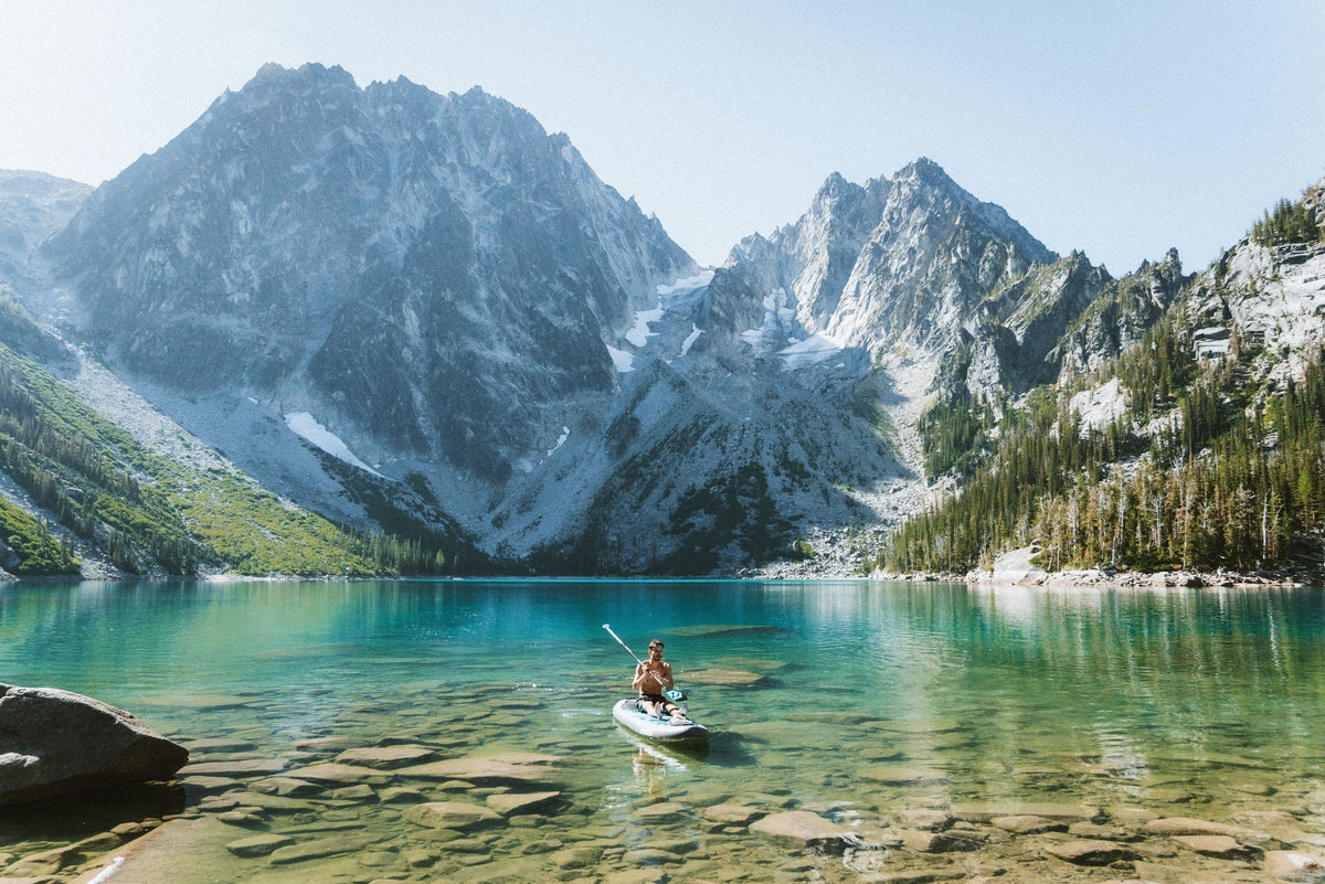 A paddler on a Hydrus inflatable paddle board crossing a clear alpine lake below rugged mountain peaks