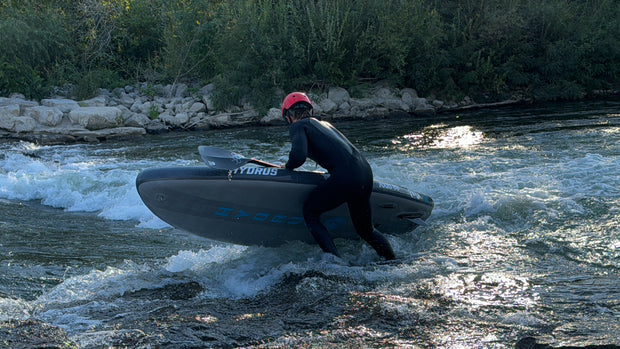 A paddler riding a Hydrus inflatable paddleboard through a whitewater river wave, spray flying off the rail