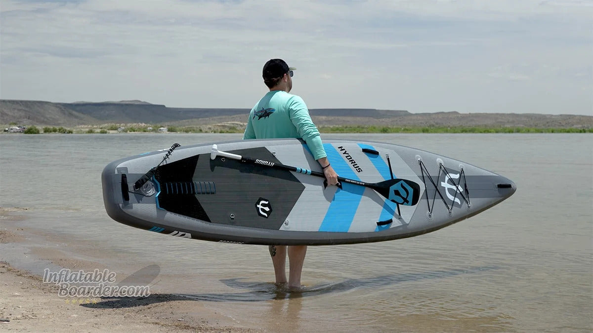 A paddler carrying a Hydrus inflatable paddleboard into a calm lake, choosing a board matched to their water