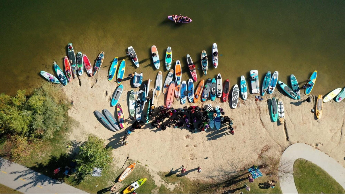Six paddleboard types arranged on a dock: all-around, touring, surfing, river/whitewater, race, and multi-person. Each shape visible in profile showing the differences in width, length, and nose shape.