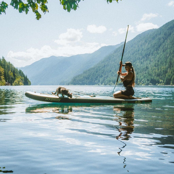 A Hydrus paddleboarder kneeling on a calm mountain lake with a cat riding the nose of the board